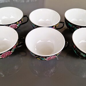 May include: Six white porcelain teacups with black handles and rims. The rims are decorated with a floral pattern in pink, yellow, and green against a black background. The cups are arranged in a row.