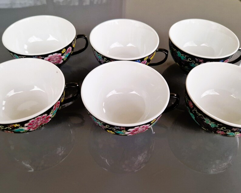 May include: Six white porcelain teacups with black handles and rims. The rims are decorated with a floral pattern in pink, yellow, and green against a black background. The cups are arranged in a row.