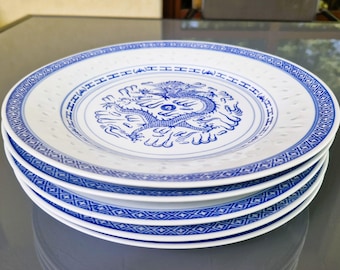 Puede incluir: Una pila de platos de cerámica blanca con una ilustración de un dragón azul en el centro. El borde de cada plato está decorado con un patrón geométrico azul y una serie de pequeñas hendiduras circulares en relieve alrededor del borde.