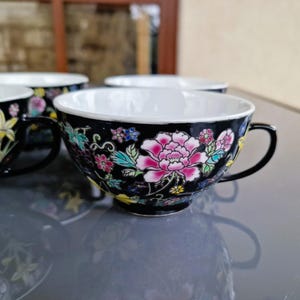May include: Set of four black teacups with white interiors and floral designs. Each cup features a large pink peony and other colorful flowers. The cups have black handles and are placed on a reflective surface.