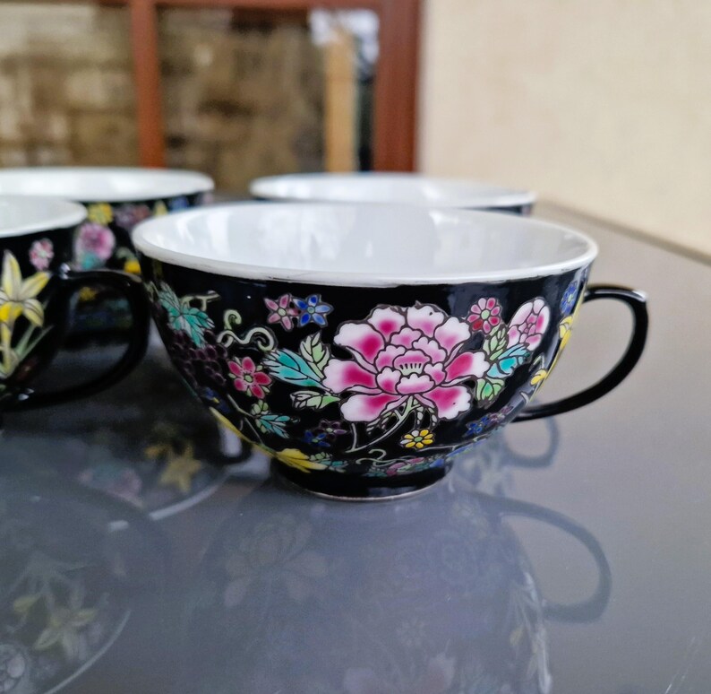 May include: Set of four black teacups with white interiors and floral designs. Each cup features a large pink peony and other colorful flowers. The cups have black handles and are placed on a reflective surface.