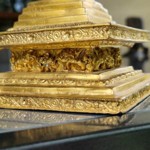 May include: Close-up of a gold-colored, tiered decorative object. The object has intricate floral and scrollwork patterns along the edges and base. The object appears to be made of metal and has a reflective surface.