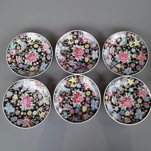May include: Six round decorative plates with a black background and a floral design. The plates feature a central pink flower surrounded by smaller flowers in various colors, including blue, yellow, and white. Each plate has a white rim.
