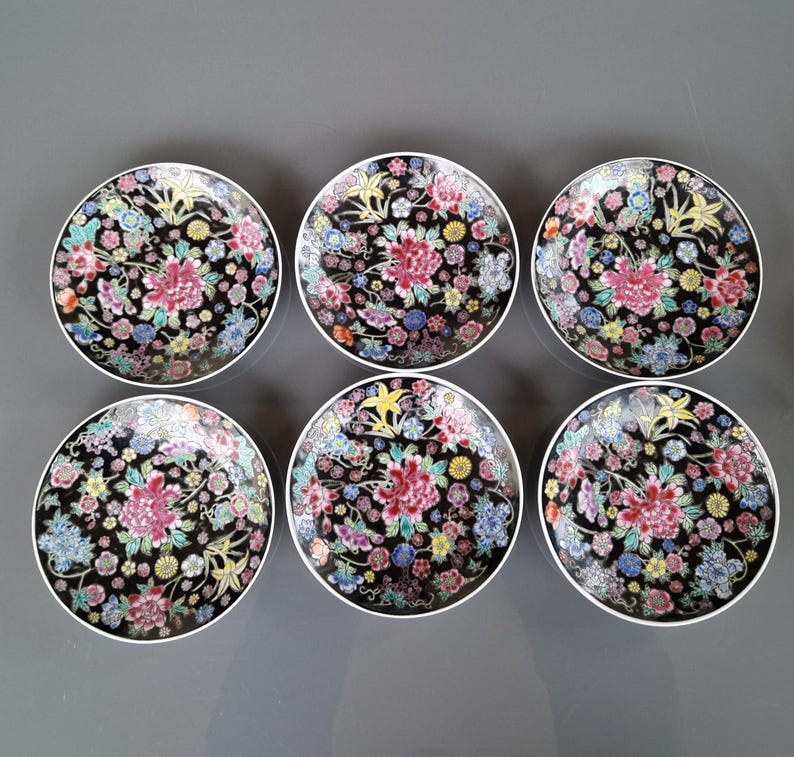 May include: Six round decorative plates with a black background and a floral design. The plates feature a central pink flower surrounded by smaller flowers in various colors, including blue, yellow, and white. Each plate has a white rim.