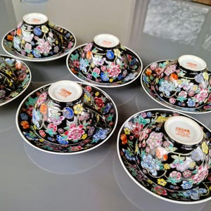 May include: Set of six black teacups and saucers with a floral design in pink, blue, yellow, and white. The cups and saucers have a white rim and a glossy finish. The teacups have a handle.