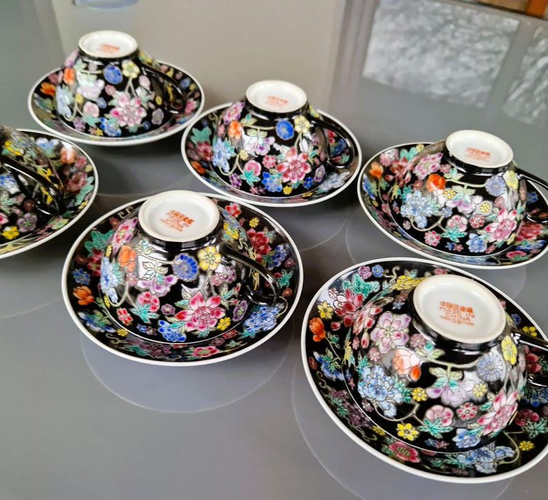May include: Set of six black teacups and saucers with a floral design in pink, blue, yellow, and white. The cups and saucers have a white rim and a glossy finish. The teacups have a handle.