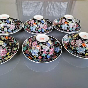May include: Six black teacups and saucers with floral designs. The cups and saucers have a black background with colorful flowers in pink, blue, yellow, and orange. The cups have black handles and white rims.