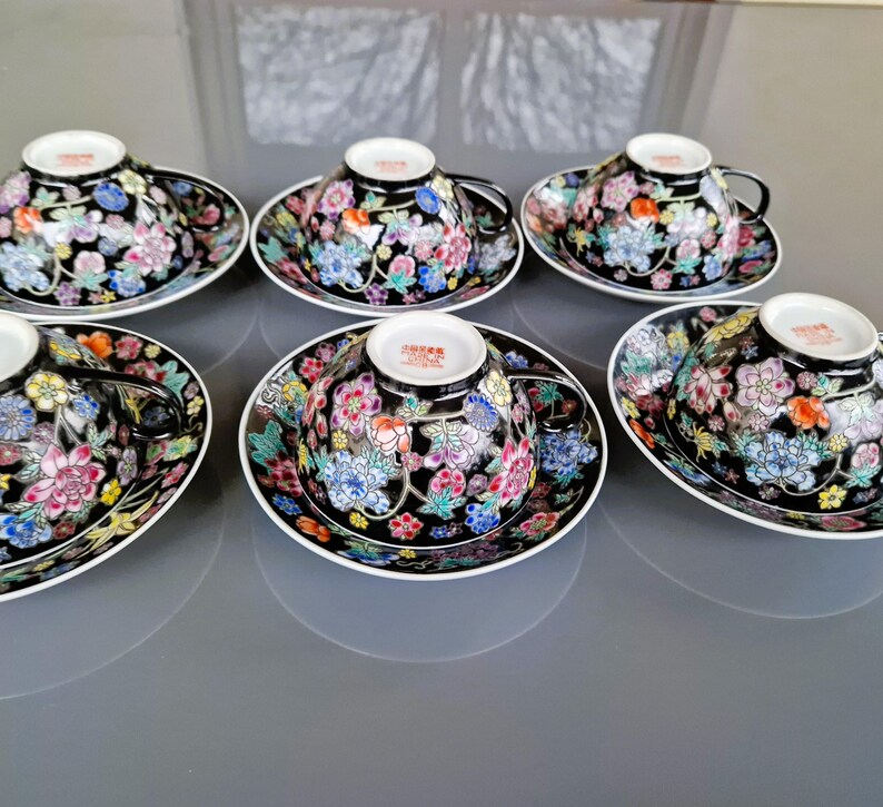 May include: Six black teacups and saucers with floral designs. The cups and saucers have a black background with colorful flowers in pink, blue, yellow, and orange. The cups have black handles and white rims.