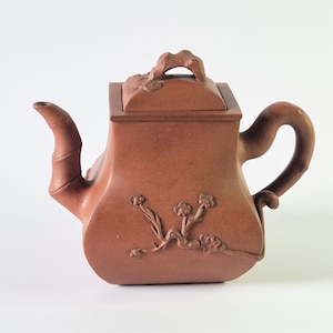 May include: A terracotta teapot with a square body and a bamboo-shaped spout. The teapot has a lid with a carved design and a floral motif on the side.