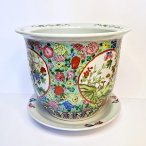 May include: A white porcelain planter with a saucer. The planter is decorated with a floral pattern in shades of pink, red, green, blue, and yellow. The planter has a round shape and a wide rim.