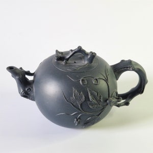 以下が含まれることがあります： 丸いボディとブドウの模様が施された黒いティーポット。ティーポットは枝のハンドルと枝の蓋が付いています。