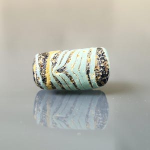 May include: A cylindrical glass bead with a striped pattern in light blue, yellow, and black. The bead's surface has a textured appearance, and its reflection is visible below. This decorative bead could be used for jewellery making or other crafts.