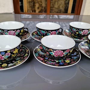May include: Set of six black teacups and saucers with a floral design in pink, yellow, blue, and green. The teacups have white interiors and black handles. The saucers match the teacups with the same floral pattern.