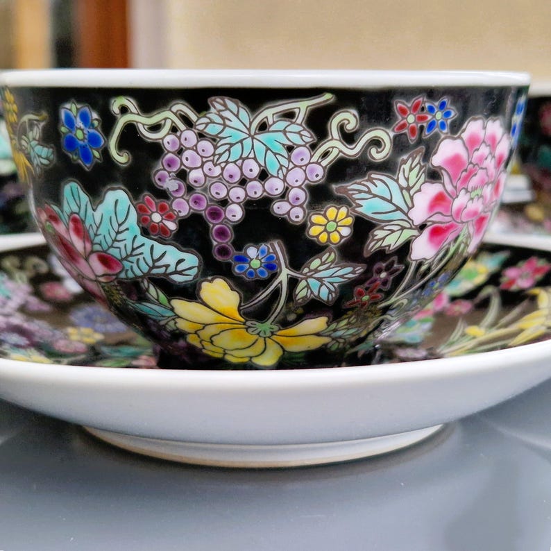 May include: A black teacup and saucer set with a white rim. The teacup and saucer are decorated with colorful floral and grape vine designs. The flowers are pink, yellow, blue, and red. The teacup is sitting on a white saucer.