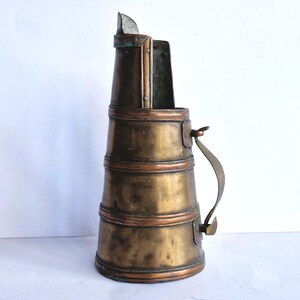 May include: A tall, tapered copper vessel with a weathered, metallic patina. The container features three horizontal raised bands, a curved metal handle attached to the side, and an open, notched top rim with a vertical spout-like extension.