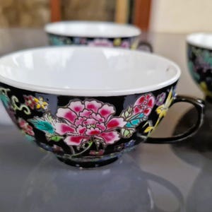 May include: A set of black teacups with a floral design. The cups have a white interior and are decorated with pink, red, and teal flowers. The handles are black.