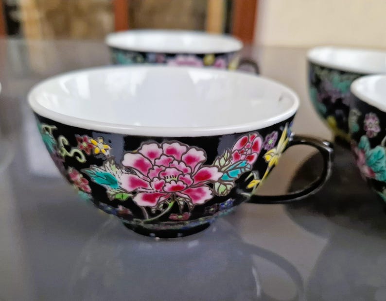 May include: A set of black teacups with a floral design. The cups have a white interior and are decorated with pink, red, and teal flowers. The handles are black.
