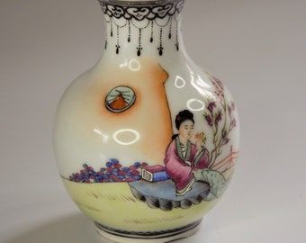 May include: A small, white porcelain vase with a round body and a narrow neck. The vase is decorated with a hand-painted scene of a woman in a pink dress sitting on a rock, surrounded by flowers and a bridge. The vase has a black and white border around the top.