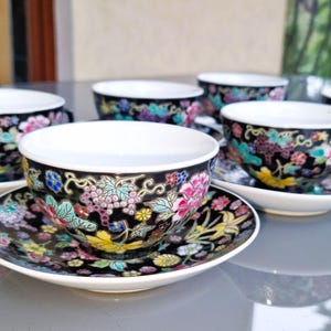 May include: Set of six porcelain teacups and saucers. The cups and saucers are black with a floral design in pink, purple, yellow, and teal. The cups have white interiors and small handles. The saucers are round and have a matching floral pattern.