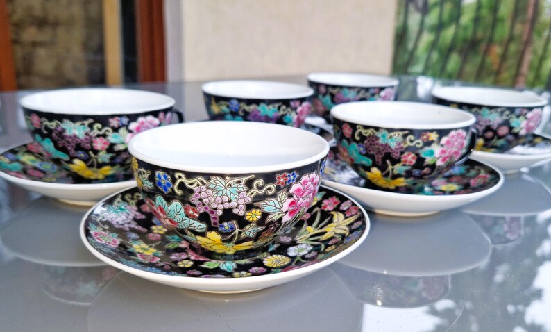 May include: Set of six porcelain teacups and saucers. The cups and saucers are black with a floral design in pink, purple, yellow, and teal. The cups have white interiors and small handles. The saucers are round and have a matching floral pattern.
