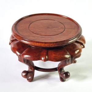 May include: A dark brown wooden pedestal with a round top and three legs. The pedestal has a carved design around the top and bottom.