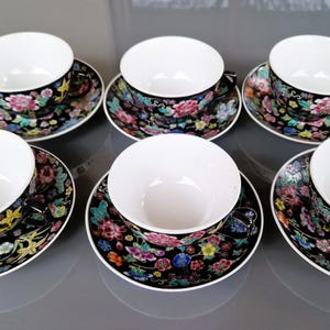 May include: Six matching teacups and saucers. The cups and saucers have a black background with a floral pattern in pink, yellow, blue, and green. The cups are white inside.