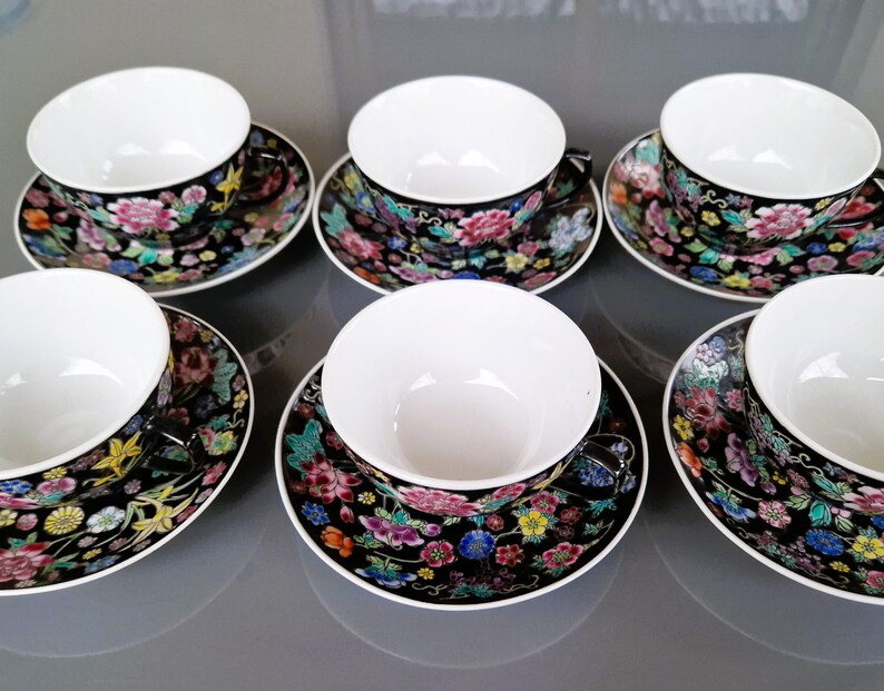 May include: Six matching teacups and saucers. The cups and saucers have a black background with a floral pattern in pink, yellow, blue, and green. The cups are white inside.
