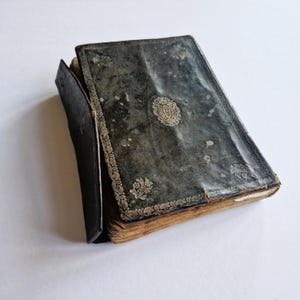 以下が含まれることがあります： 金色のアクセントが付いた、黒革製のアンティークな本。古びていて、使い込まれたような外観です。本は閉じられており、白い表面の上に置かれています。