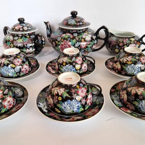 May include: A black tea set with floral patterns. The set includes a teapot, a creamer, and several teacups with saucers. The floral design features pink, blue, and white flowers. The set is arranged on a white surface.