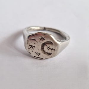 May include: A silver ring with a rectangular face. The face has etched designs including a crescent moon, stars, and the letters "I" and "K". The ring has a slightly worn appearance.