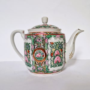 May include: A white porcelain teapot with a green, pink, and gold floral pattern. The teapot has a round body and a spout on the right side.