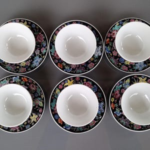 May include: Six white teacups sit on matching saucers. The saucers have a black background with a colorful floral pattern. The teacups are round and have a simple, elegant design. The set is arranged on a gray surface.