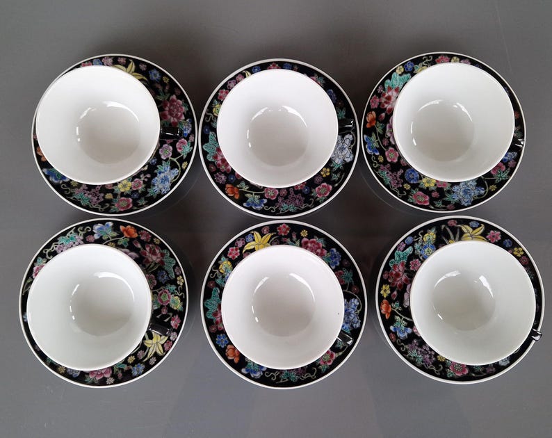 May include: Six white teacups sit on matching saucers. The saucers have a black background with a colorful floral pattern. The teacups are round and have a simple, elegant design. The set is arranged on a gray surface.