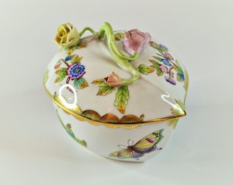 May include: A white porcelain trinket box with a floral design and a butterfly on the bottom. The lid has a green stem with a pink and yellow flower on top.