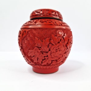 Könnte beinhalten: Eine kleine, rote, kunstvoll geschnitzte chinesische Zinnoberlackdose mit floralem Design. Die Dose hat einen Deckel und ist mit einem sich wiederholenden Muster aus Blumen und Blättern verziert.