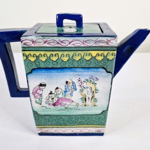 以下が含まれることがあります： 青い取っ手、注ぎ口、蓋の取っ手が付いた正方形のセラミックティーポット。本体は、緑、黄、ピンクのアクセントで、風景の中の人物のシーンで装飾されています。ティーポットは正方形です。
