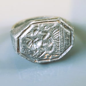 May include: A silver-toned ring with an octagonal face. The face features an engraved design of a bird or mythical creature. The ring has a worn, antique appearance, suggesting age and history. The ring is a piece of jewelry.