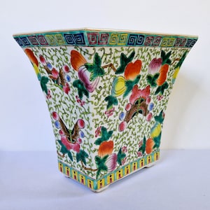 Könnte beinhalten: Ein quadratischer, weißer Keramik-Blumentopf mit einem farbenfrohen Blumen- und Schmetterlingsdesign. Der Pflanztopf hat einen geometrischen Rand und ist mit roten, rosa, gelben und grünen Elementen verziert. Der Pflanztopf hat eine leicht ausgestellte Form und steht auf kleinen Füßen.