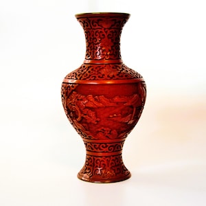 Peut inclure: Un vase rouge profond de style cinabre orné de sculptures en relief complexes représentant un paysage avec des arbres et des montagnes. Le col et la base présentent des motifs de volutes détaillés, tandis que le corps central affiche un relief scénique.