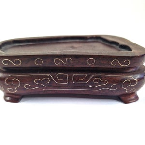 May include: A dark brown wooden bonsai pot with intricate gold and white inlay designs. The pot has four legs and a rectangular shape.