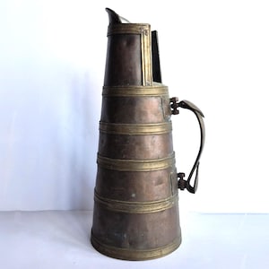 May include: An antique copper pitcher with brass accents. The pitcher has a tall, cylindrical body with horizontal brass bands and a hinged handle. The spout is slightly curved, and the overall design suggests a vintage aesthetic.