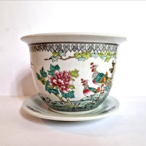May include: A white ceramic planter with a matching saucer, featuring a hand-painted design of colourful roosters, vibrant pink peonies, and green foliage. The rim is decorated with a black and white geometric pattern, and the base is flared.