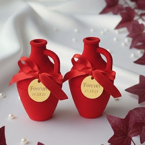 Puede incluir: Dos pequeños jarrones rojos con asas y lazos rojos, cada uno con una etiqueta dorada que dice "Forever 11.13.25". Los jarrones están rodeados de hiedra burdeos y perlas, sobre un fondo blanco.