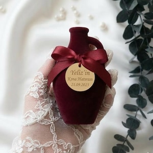 Può includere: Un piccolo vaso bordeaux con manico, ornato da un nastro abbinato e un'etichetta dorata. L'etichetta recita "Yeliz'in Kına Hatırası 21.09.2025". Il vaso è tenuto da una persona con guanti di pizzo bianco.