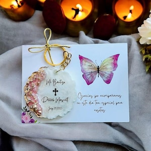 May include: White and gold round keychain with a cross and the text "Mi Bautizo Delilah Maywatt July 29, 2023". A pink and white butterfly is painted on a white card with the text "Gracias por acompañamos en este dia tan especial para nosotros".