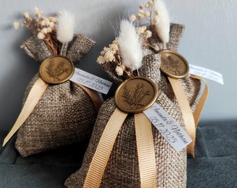 May include: Three small burlap bags with dried flowers and a gold wax seal. Each bag has a gold ribbon and a tag with the text "Amanda & Nathan 05.17.2023".