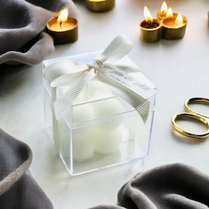 May include: A clear square box with a white ribbon and bow, containing four white candles. The box has a small white tag attached with the text "Lyse & Ale 09.09.2021".