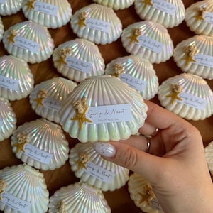 Puede incluir: Una colección de cajas blancas con forma de concha marina con detalles dorados y una pequeña estrella de mar en cada una. Las cajas están etiquetadas con el texto "Serife & Mesut Nişan Hatırası".