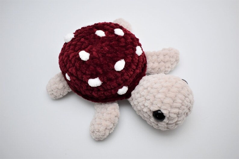 May include: A handmade plush turtle with a burgundy shell dotted with white spots. The turtle's head and limbs are a soft beige colour, and it has black eyes. The plush toy is made of a chunky, textured yarn.