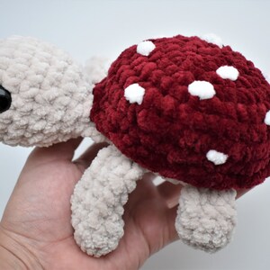 May include: A crocheted turtle toy with a mushroom-style shell. The turtle's body and limbs are beige, and the shell is a deep red with white spots. The turtle has a large, black eye, and is handmade.